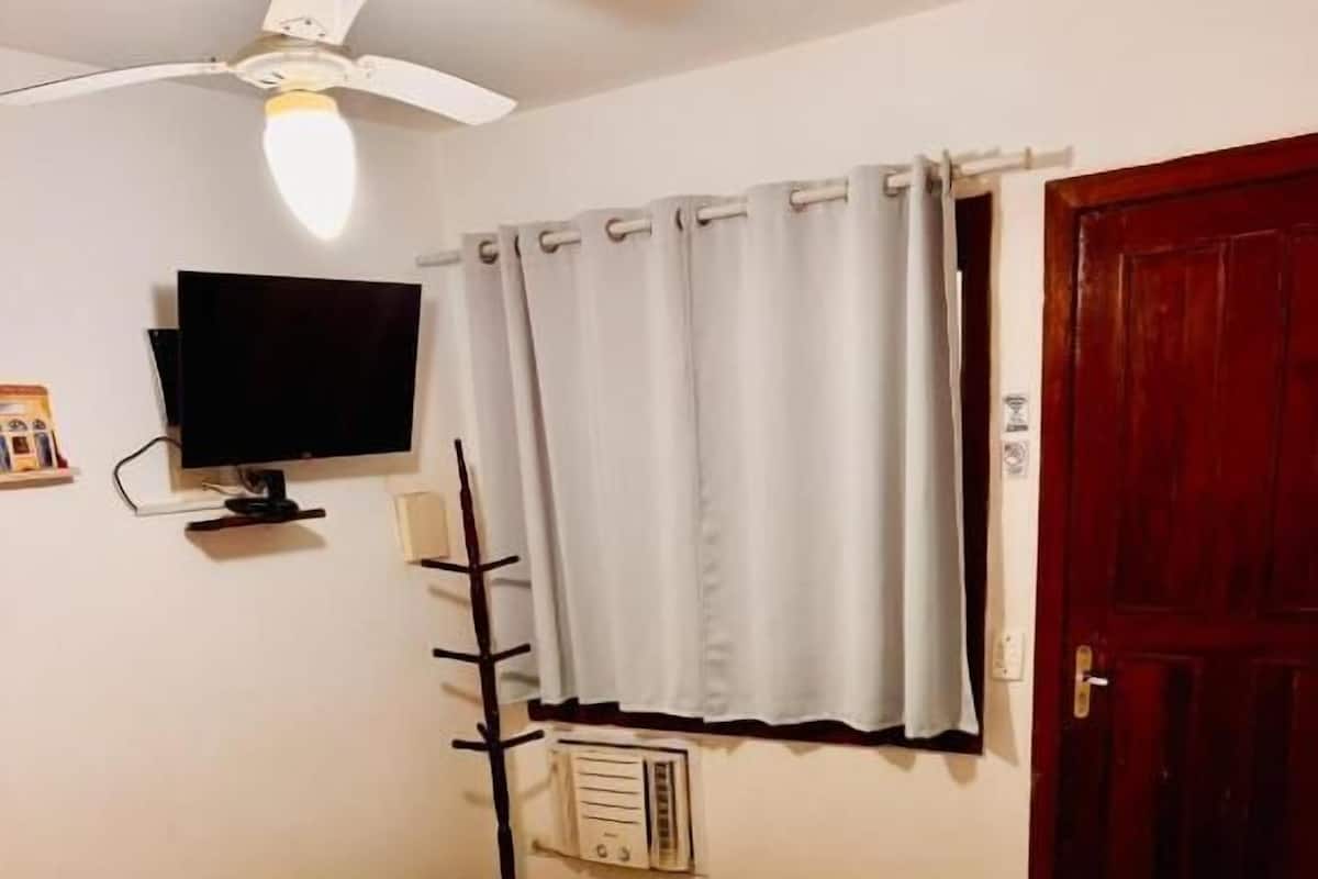 A neatly made bed with white linens, a ceiling fan, a television mounted on the wall, and a window with curtains.