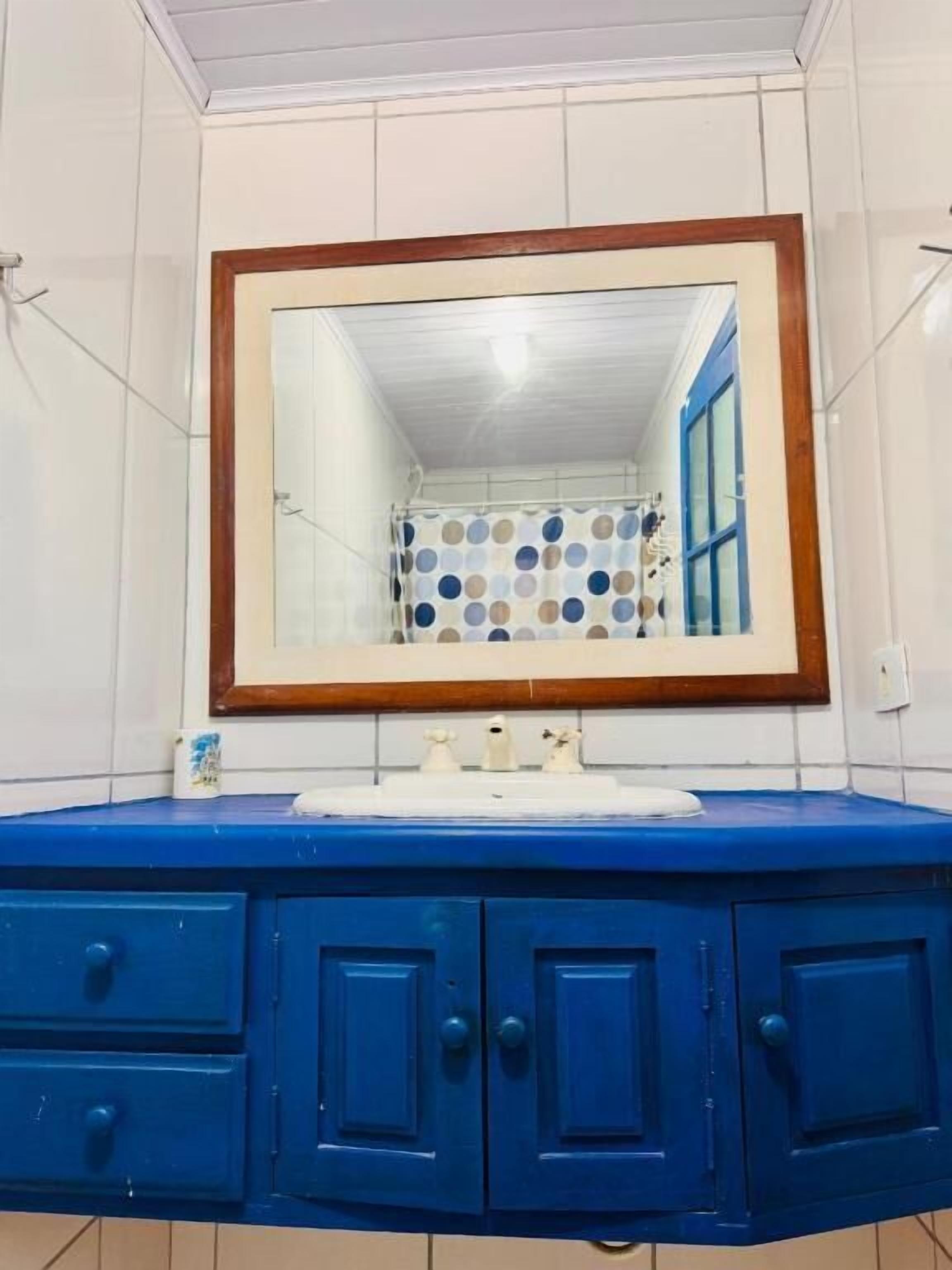 Un baño con pared de azulejos blancos, tocador azul con cajones, espejo con marco de madera y cortina de ducha a lunares.