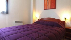 Een bed met een paars sprei, twee bedlampen, een kunstwerk aan de muur en een ventilatierooster.