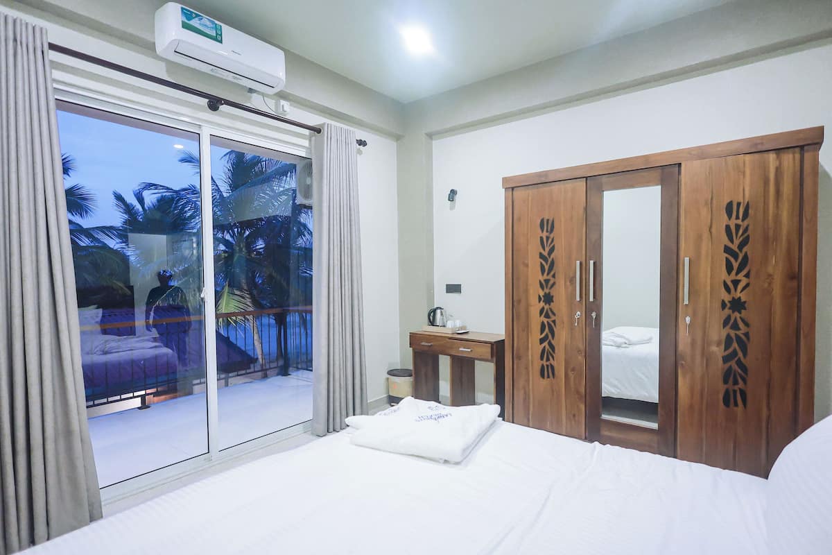 A hotel room with a bed, a wooden wardrobe, a desk with a kettle, and a balcony with a view of palm trees.