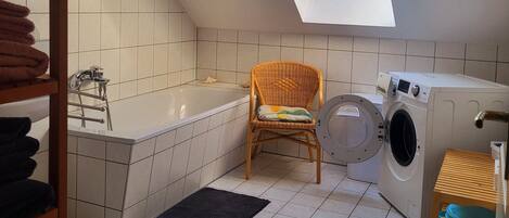 Une petite buanderie bien éclairée, dotée d’un puits de lumière, d’un lave-linge, d’une chaise et d’une baignoire.