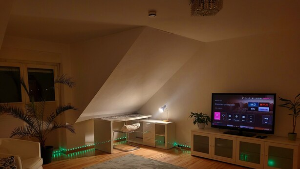 Ein gemütliches Wohnzimmer mit schrägen Wänden, einem Fernseher, einem Schreibtisch und einer Couch.