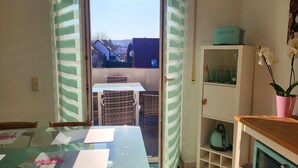 Ein Essbereich mit einem Glastisch, Korbstühlen und Blick ins Freie. Es gibt einen Schrank mit Glastüren und eine Orchidee in einem Topf auf einem Holzboden.