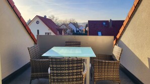 Ein kleiner Balkon mit Glastisch und Korbstühlen, umgeben von Mauern und angrenzenden Häusern.