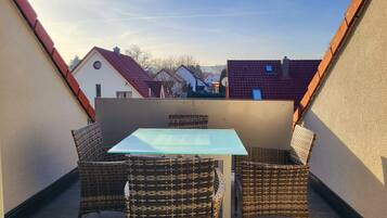 Ein kleiner Balkon mit Glastisch und Korbstühlen, umgeben von Mauern und angrenzenden Häusern.