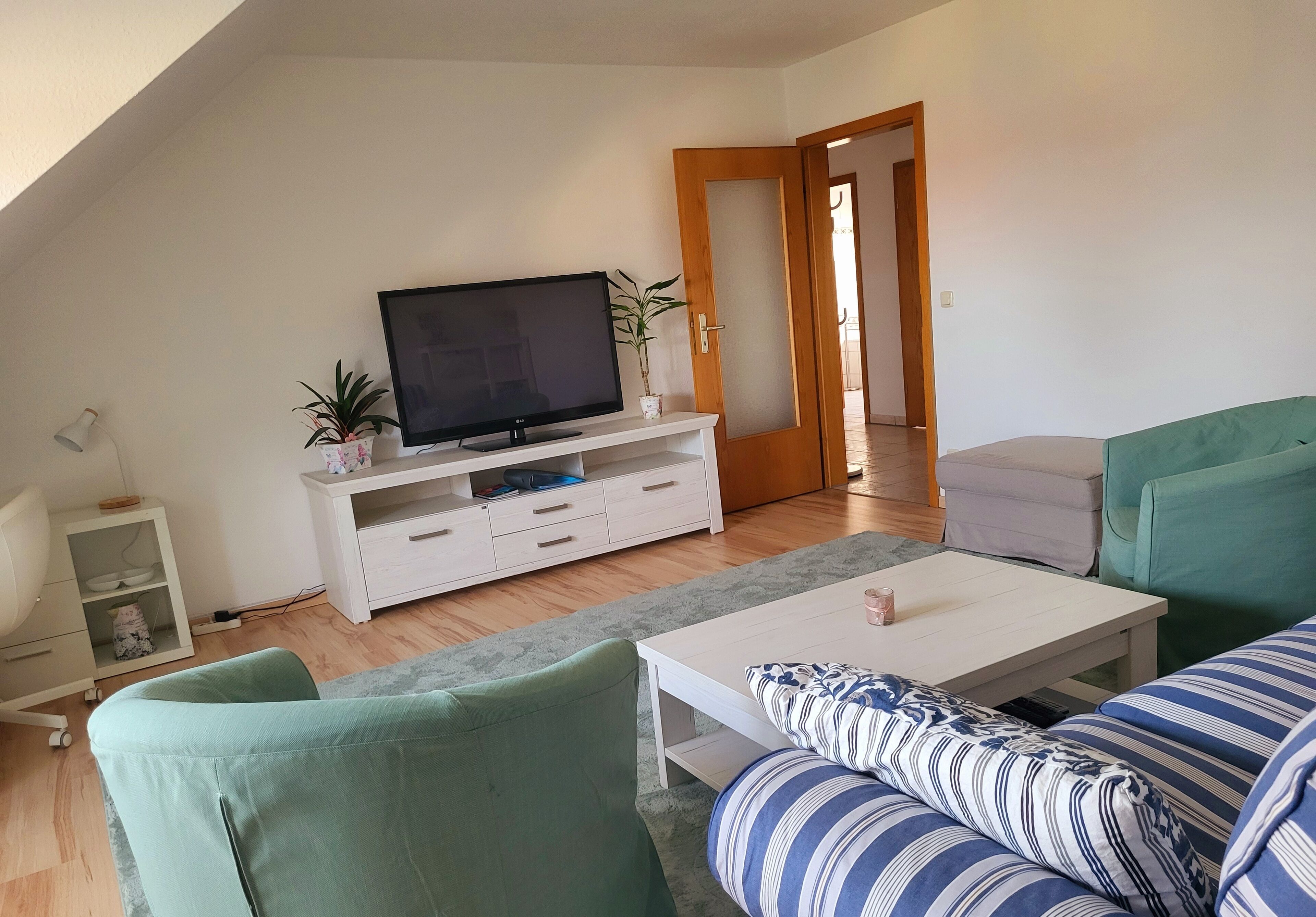 Een moderne woonkamer met een flatscreen televisie op een witte tv-meubel, een groene bank, een gestreepte bank en een witte salontafel.