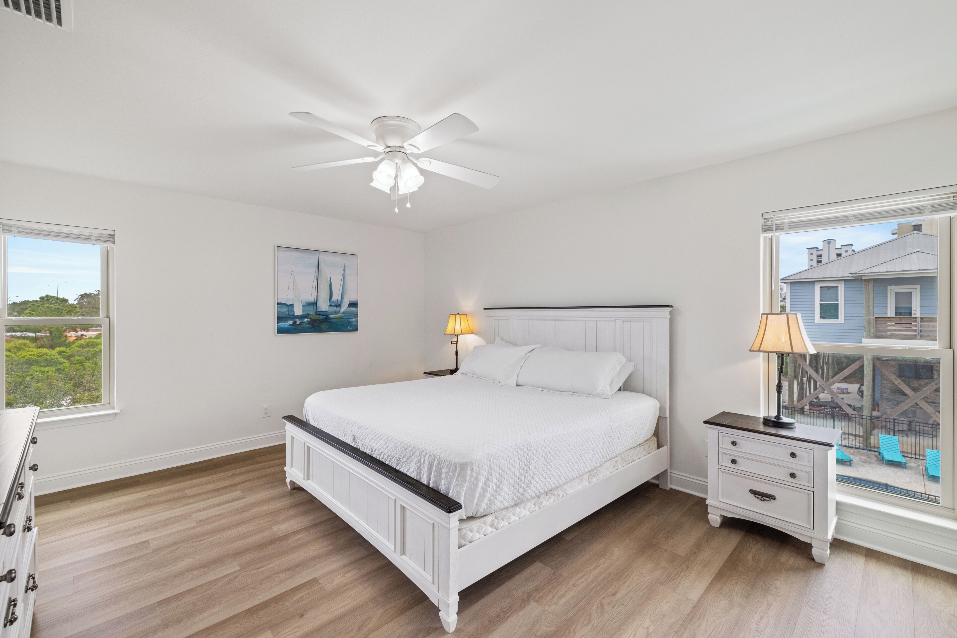 A bedroom with a white bed, a ceiling fan, a nightstand with a lamp, a window with a view of a house and greenery.