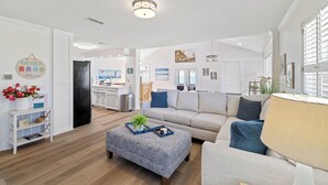 Un salon moderne avec un canapé, un pouf et une lampe. On aperçoit un coin cuisine au fond.