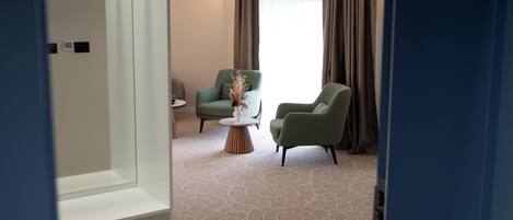 A hotel room with a carpeted floor, two green armchairs, a small round table with a vase, and a window with curtains.