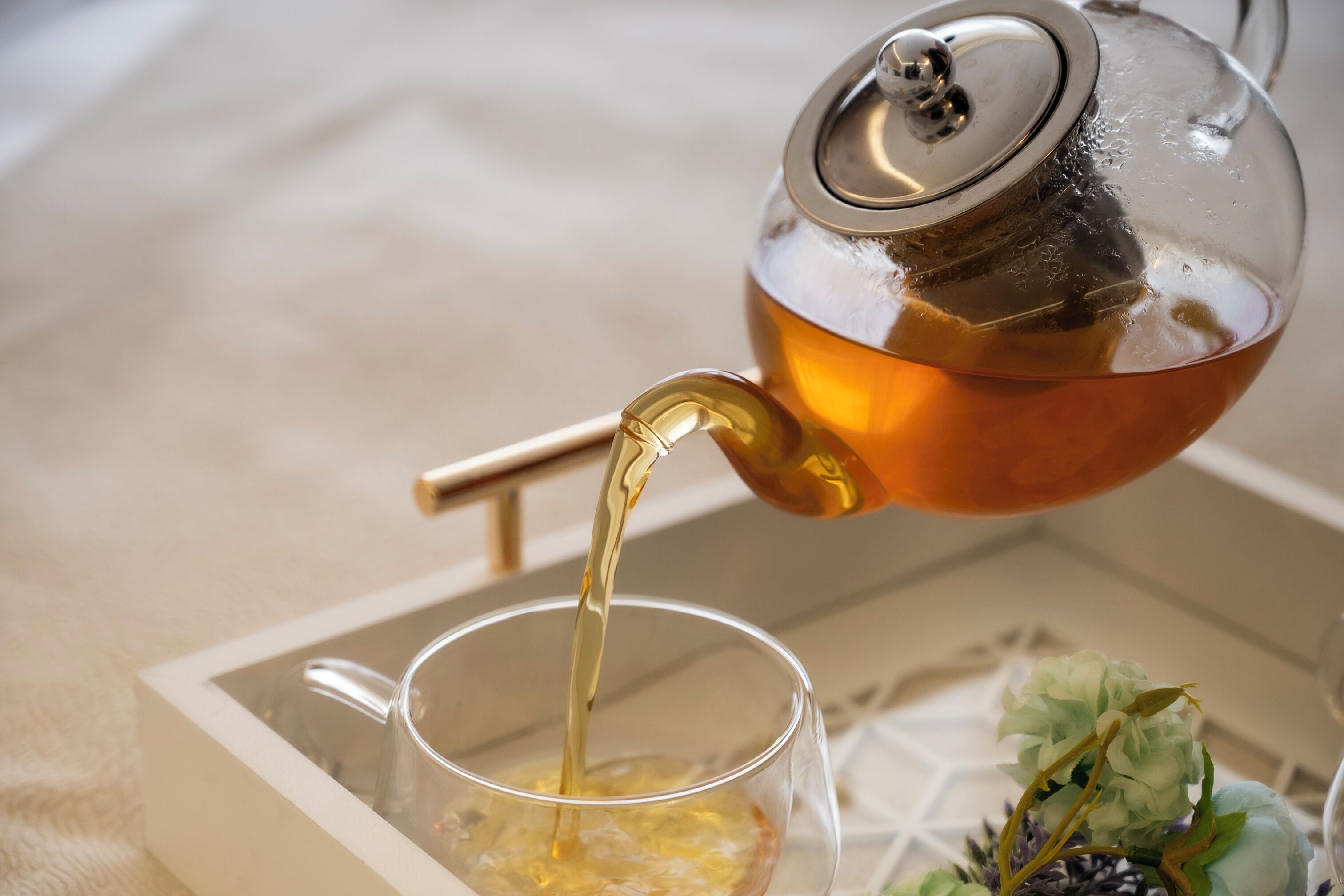 A clear glass teapot pouring tea into a glass cup on a tray.