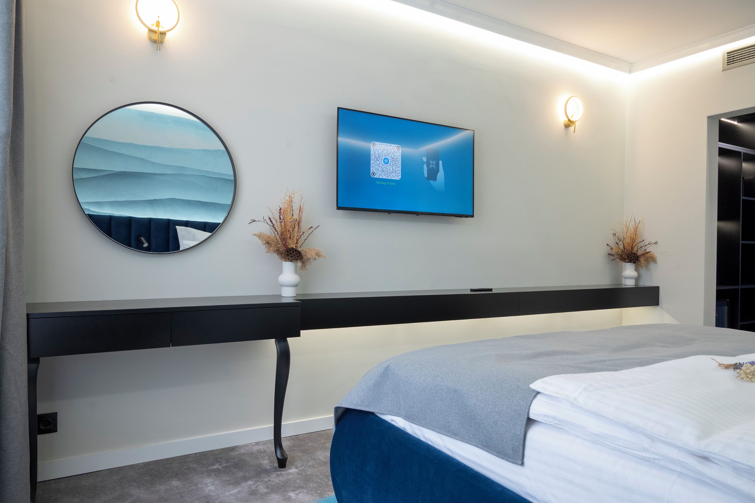 A modern bedroom with a bed, a desk, a round mirror, and a wall-mounted TV.