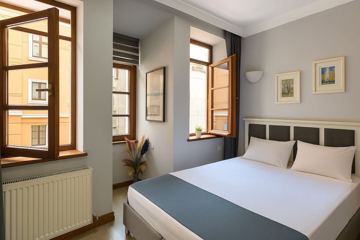 Hotellihuone, jossa on sänky, patteri, ikkuna, jonka sälekaihtimet ovat auki, ja seinälle kiinnitetty valokuvakehys.