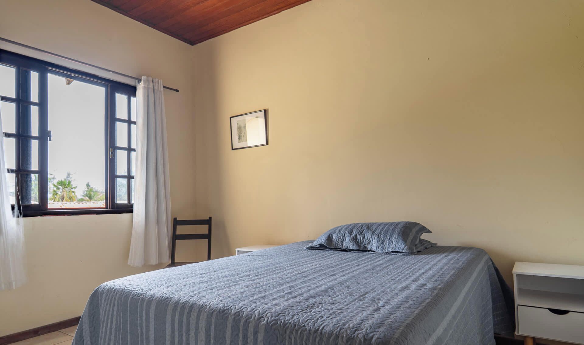 A bedroom with a bed, a chair, a window with curtains, and a framed picture on the wall.