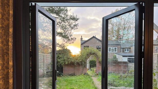 Une vue à travers une porte vitrée donnant sur un espace extérieur verdoyant, avec un bâtiment en briques en arrière-plan.
