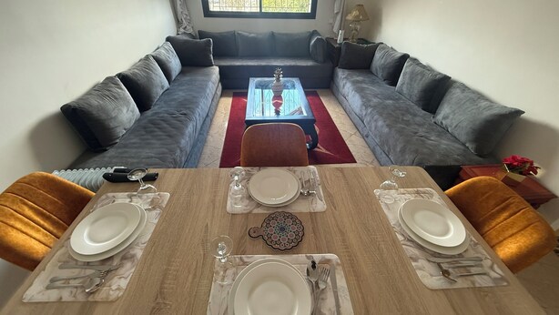 Comedor con mesa de madera para cuatro personas, dos sofás grises frente a una mesa de centro de vidrio, y una ventana con cortinas.