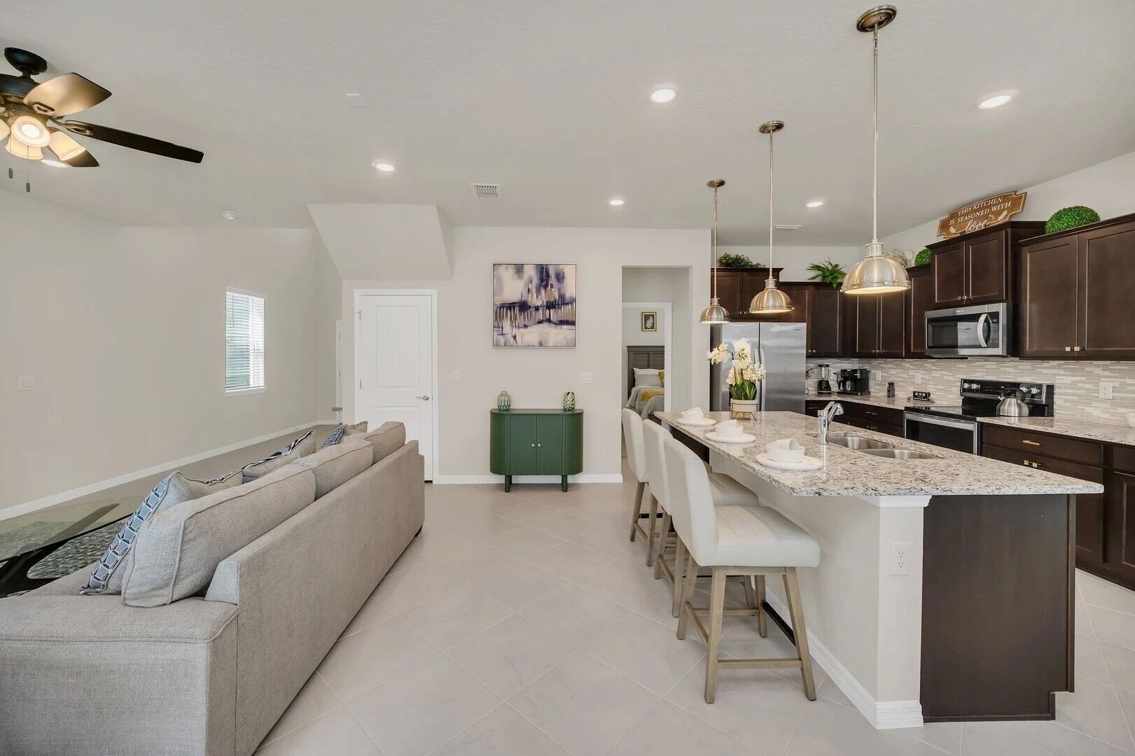 Una cocina moderna con armarios oscuros, encimera de granito y electrodomésticos de acero inoxidable. Hay una zona de comedor con un mueble verde y un cuadro en la pared.