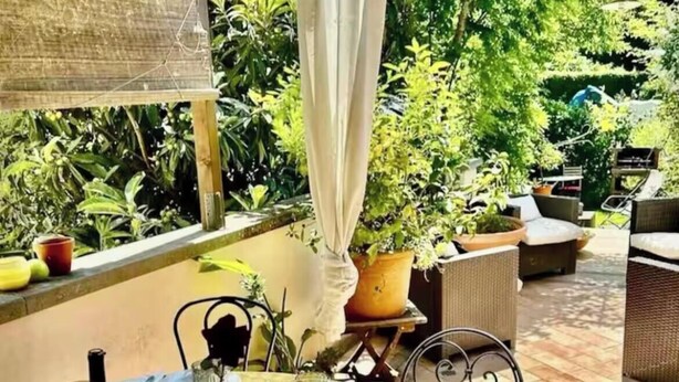 Un espace de restauration en plein air avec une table dressée pour deux personnes, entouré de plantes en pot et de meubles en osier.