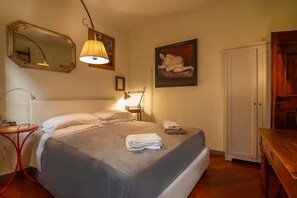 A neatly made bed with a grey bedspread and white pillows, a bedside lamp, a mirror, a painting, and a wooden wardrobe. - Charming Balcony Studio in Florence Near the City Center  (Firenze)