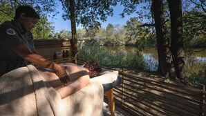 A person receiving a massage on a bench by a lake, with trees and a wooden deck in the background. - Mozhi Bushcamp  (Kafue National Park)