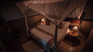 A dimly lit room with a bed under a canopy, a nightstand with a lamp, and a small rug on the floor. - Mozhi Bushcamp  (Kafue National Park)