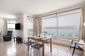 A room with a dining table, chairs, a TV, and a large window with a view of the sea and a bridge. - Apt. 'Camões 11 D' with Sea View, Wi-Fi, Air Conditioning and Pool (Armação de Pêra)