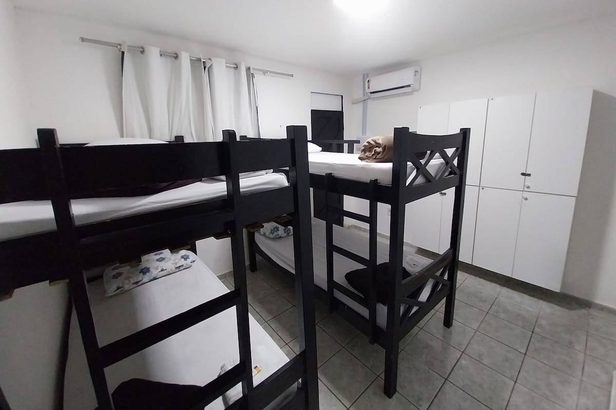 A dormitory room with bunk beds, a desk, a chair, a wardrobe, and an air conditioning unit.