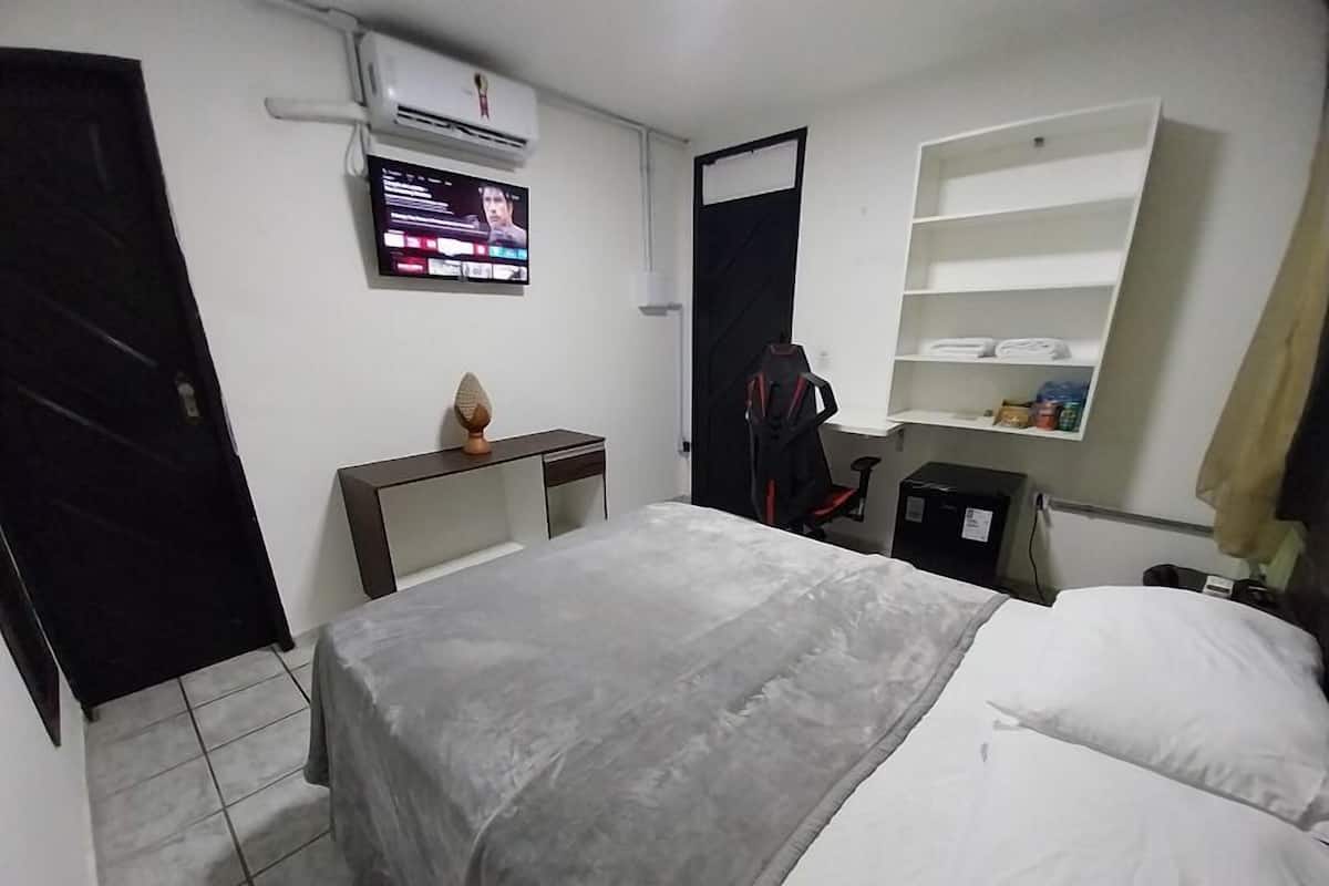 A hotel room with a bed, a desk with a chair, a TV, a wardrobe, and a shelf with toiletries.