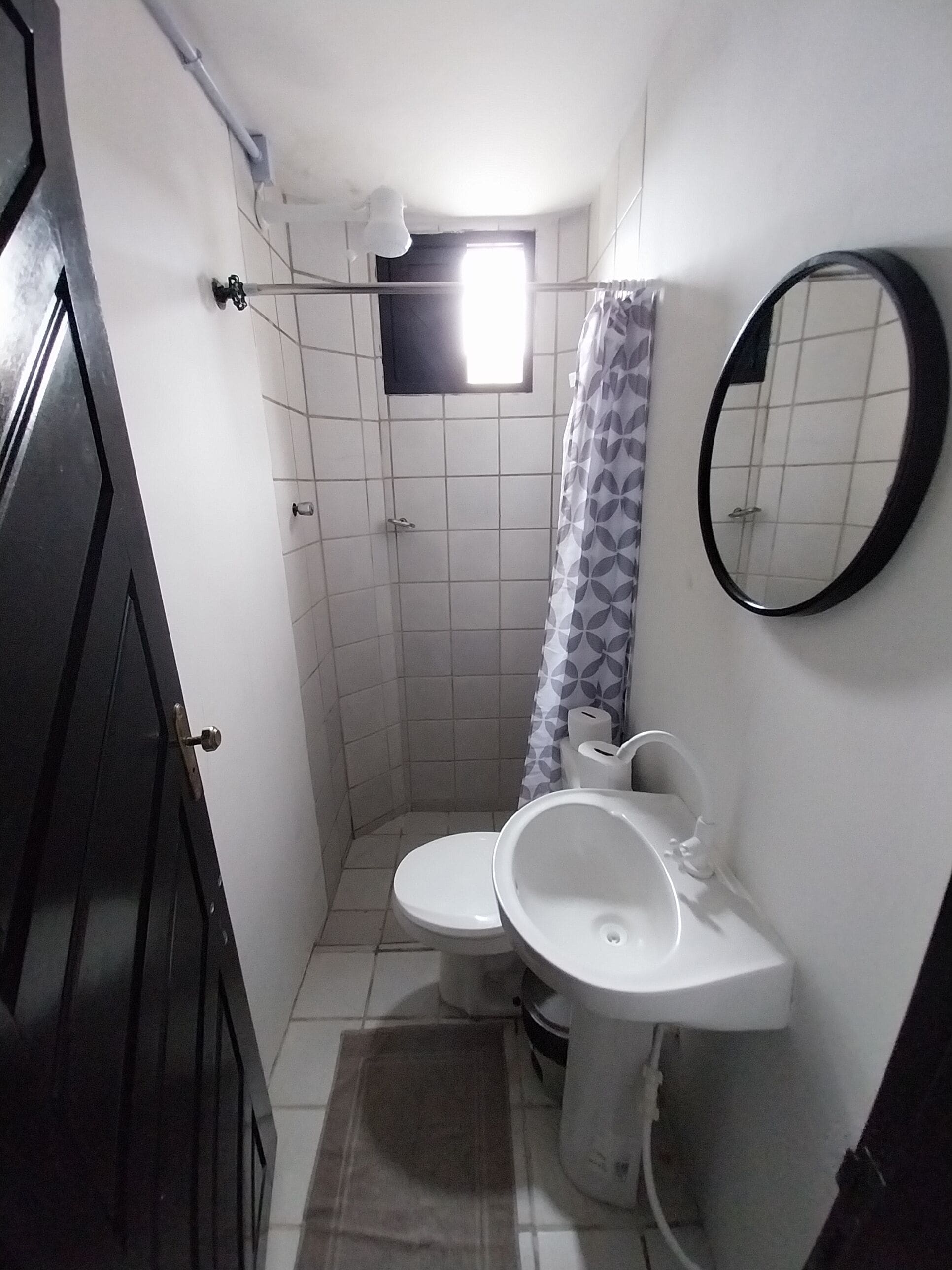 Un baño pequeño con inodoro, lavamanos y ducha. Hay una ventana sobre la ducha, un espejo en la pared y una puerta que da a otra habitación.
