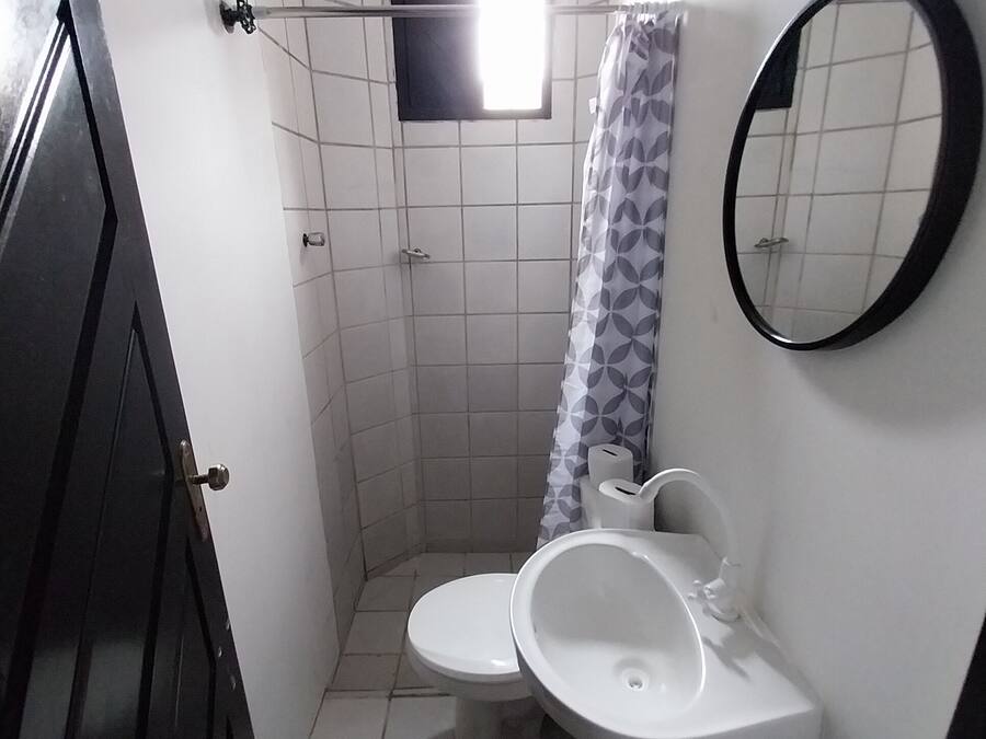 Un baño pequeño con inodoro, lavamanos y ducha. Hay una ventana sobre la ducha, un espejo en la pared y una puerta que da a otra habitación.
