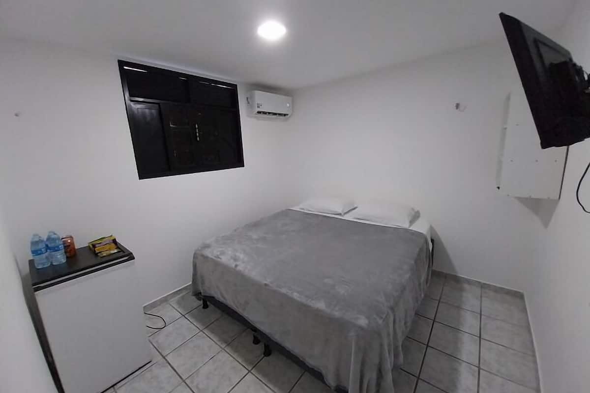 A small, clean room with a bed, a desk, a window, and an air conditioning unit.