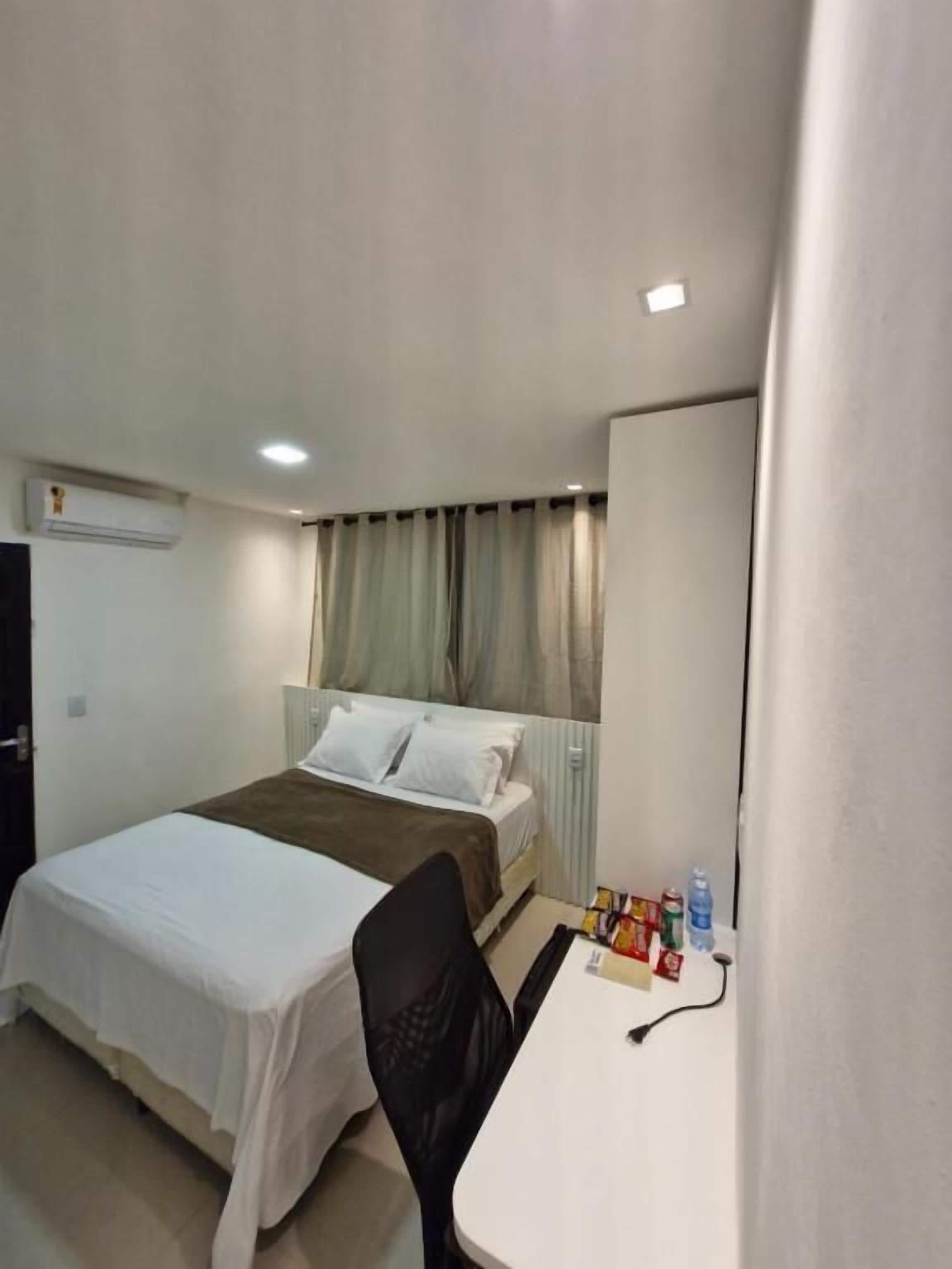 Quarto de hotel com uma cama, uma escrivaninha e uma cadeira. Há um ar condicionado na parede e uma janela com cortinas.