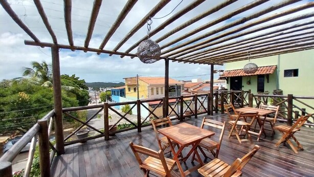 An outdoor wooden deck with tables and chairs, a bamboo pergola, and a view of buildings and greenery. - Pousada Dunas Natal (Natal)