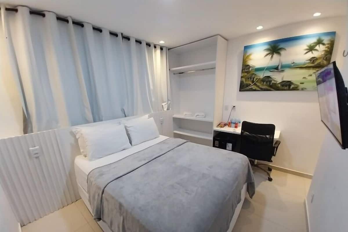 A hotel room with a bed, a desk, a chair, a painting, and a radiator.