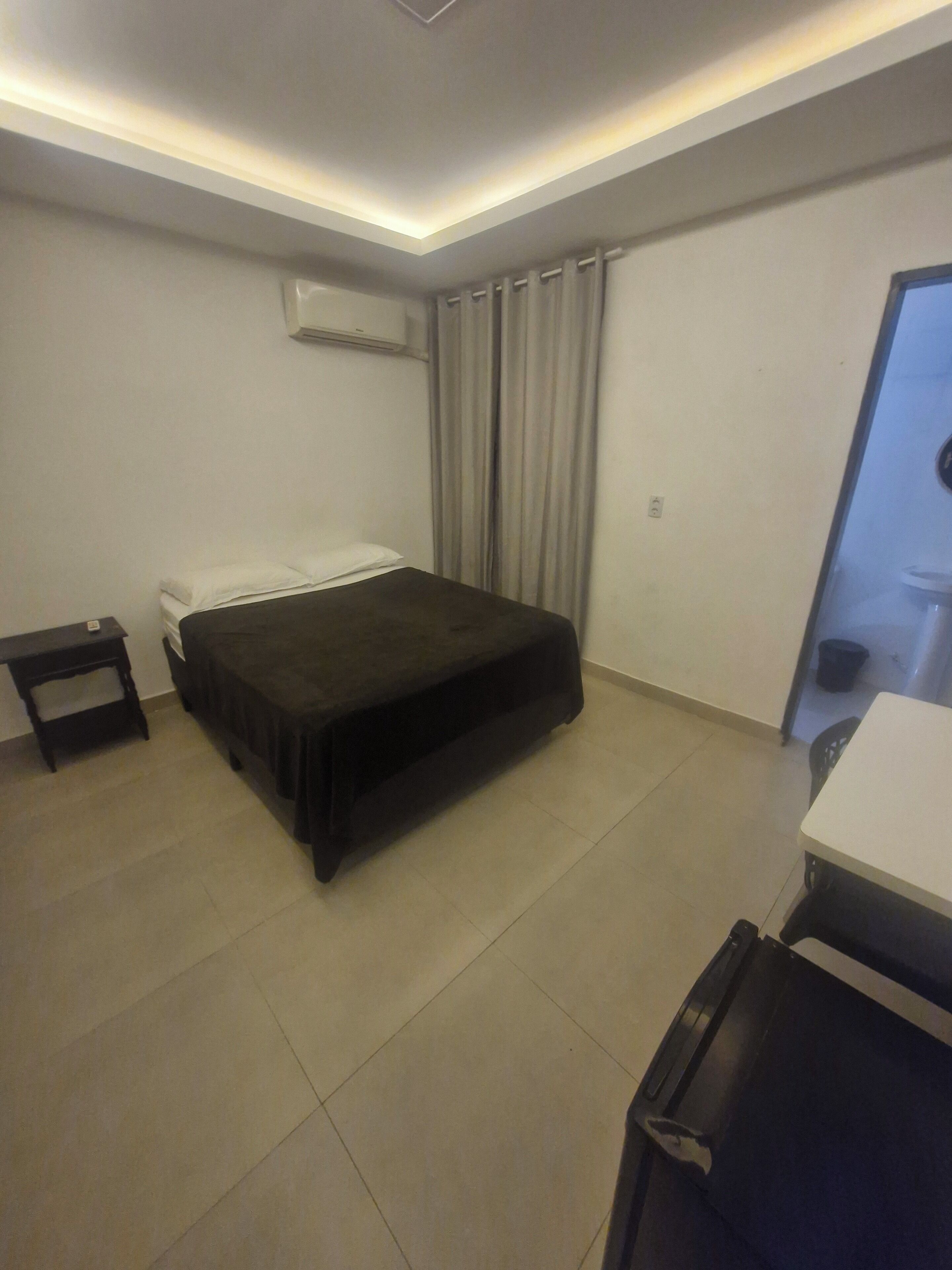 A hotel room with a bed, a small table, an air conditioning unit, a curtain, and a door.