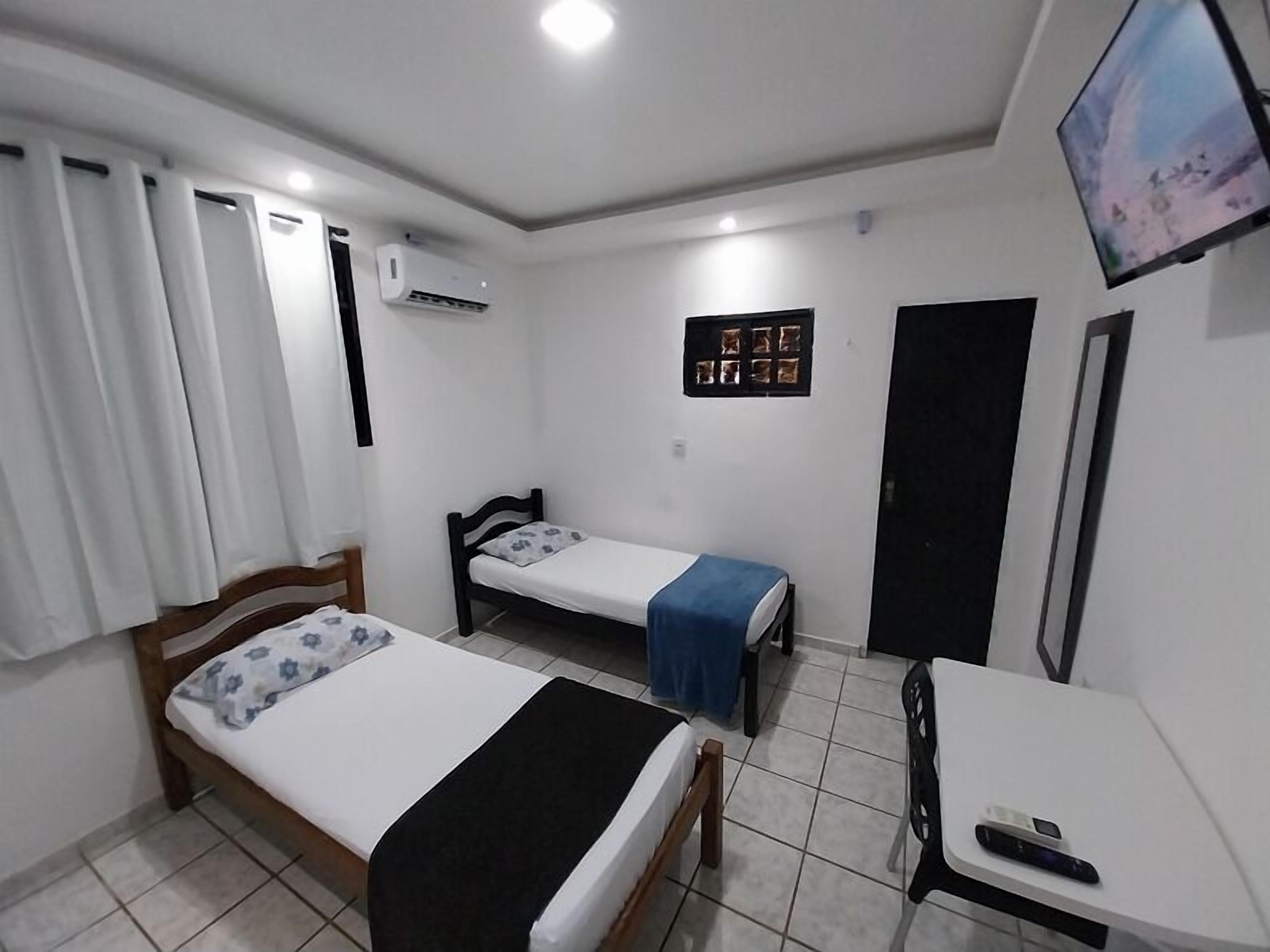 Quarto pequeno e limpo com duas camas de solteiro, uma escrivaninha, uma cadeira, uma televisão e ar condicionado.