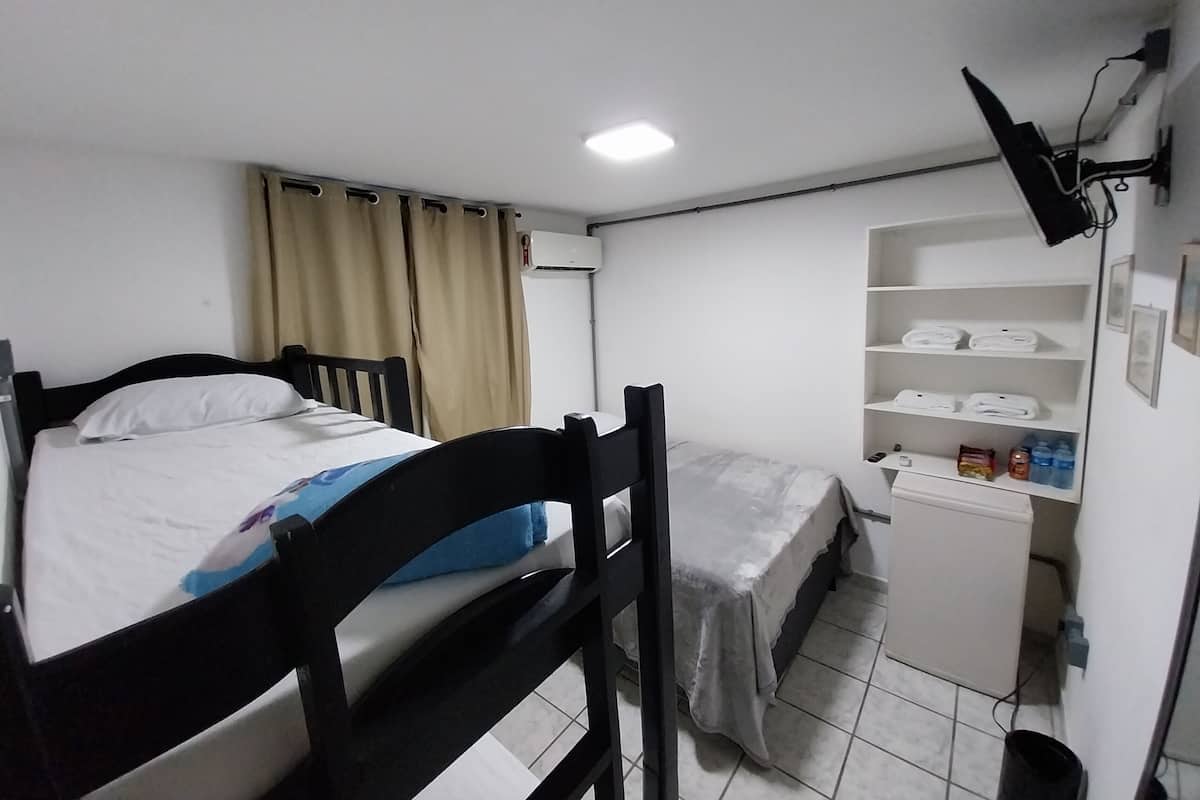 A small, clean room with two bunk beds, a desk with a chair, a shelf with toiletries, and a flat-screen TV mounted on the wall.