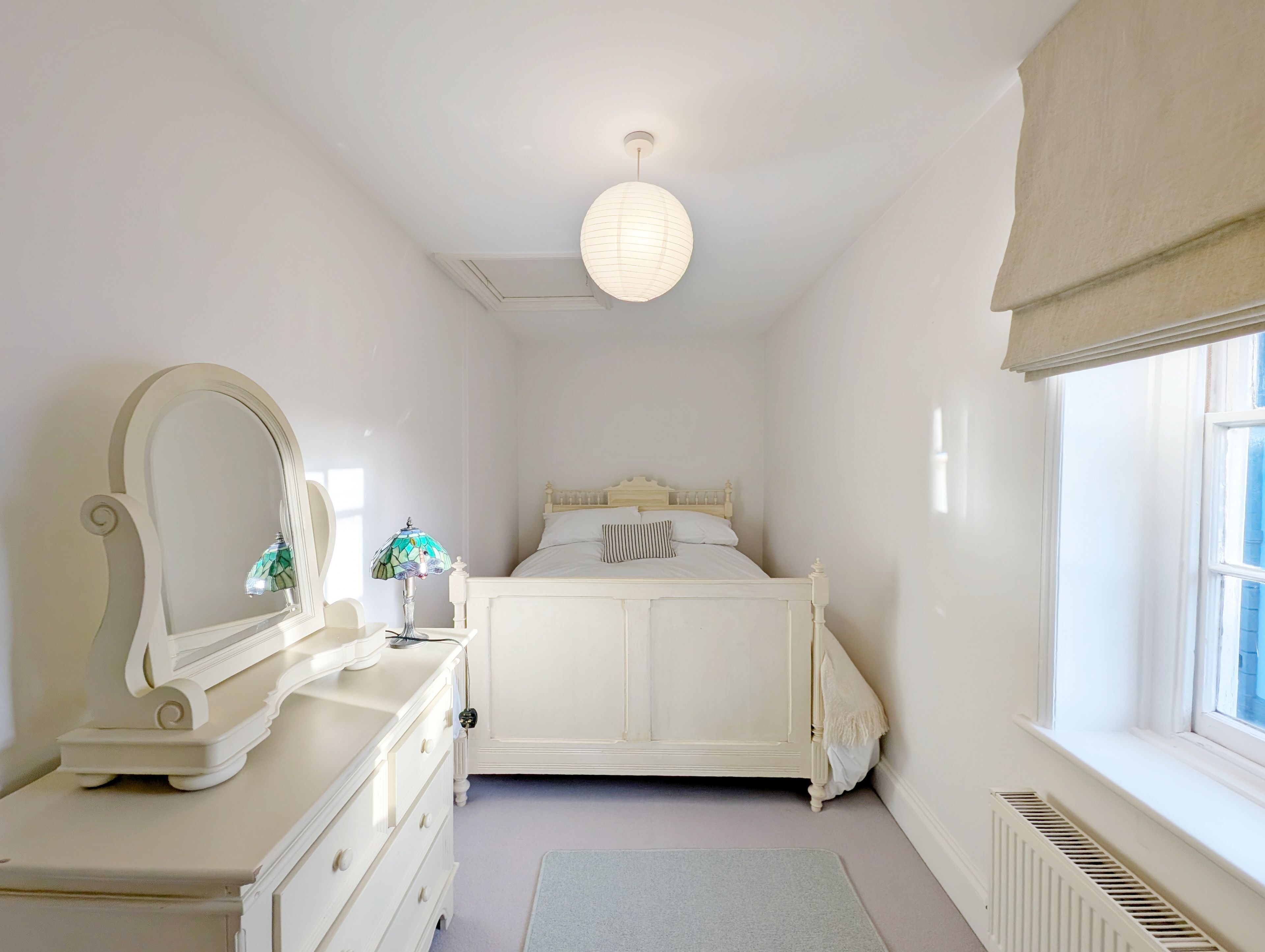 Een slaapkamer met een wit bed, een dressoir met spiegel, een hanglamp en een raam met uitzicht.