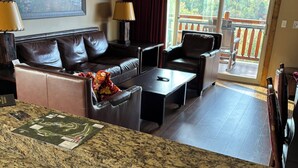 A living room with a brown leather sofa, a coffee table, and a TV. There is a painting on the wall and a window with a view of trees.