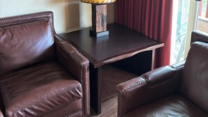 A room with two leather armchairs, a wooden table with a lamp, red curtains, and a framed picture on the wall.