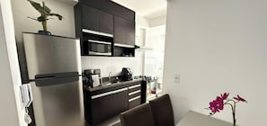 Una cocina moderna con gabinetes oscuros, electrodomésticos de acero inoxidable y un pequeño comedor con una silla y una planta en maceta.
