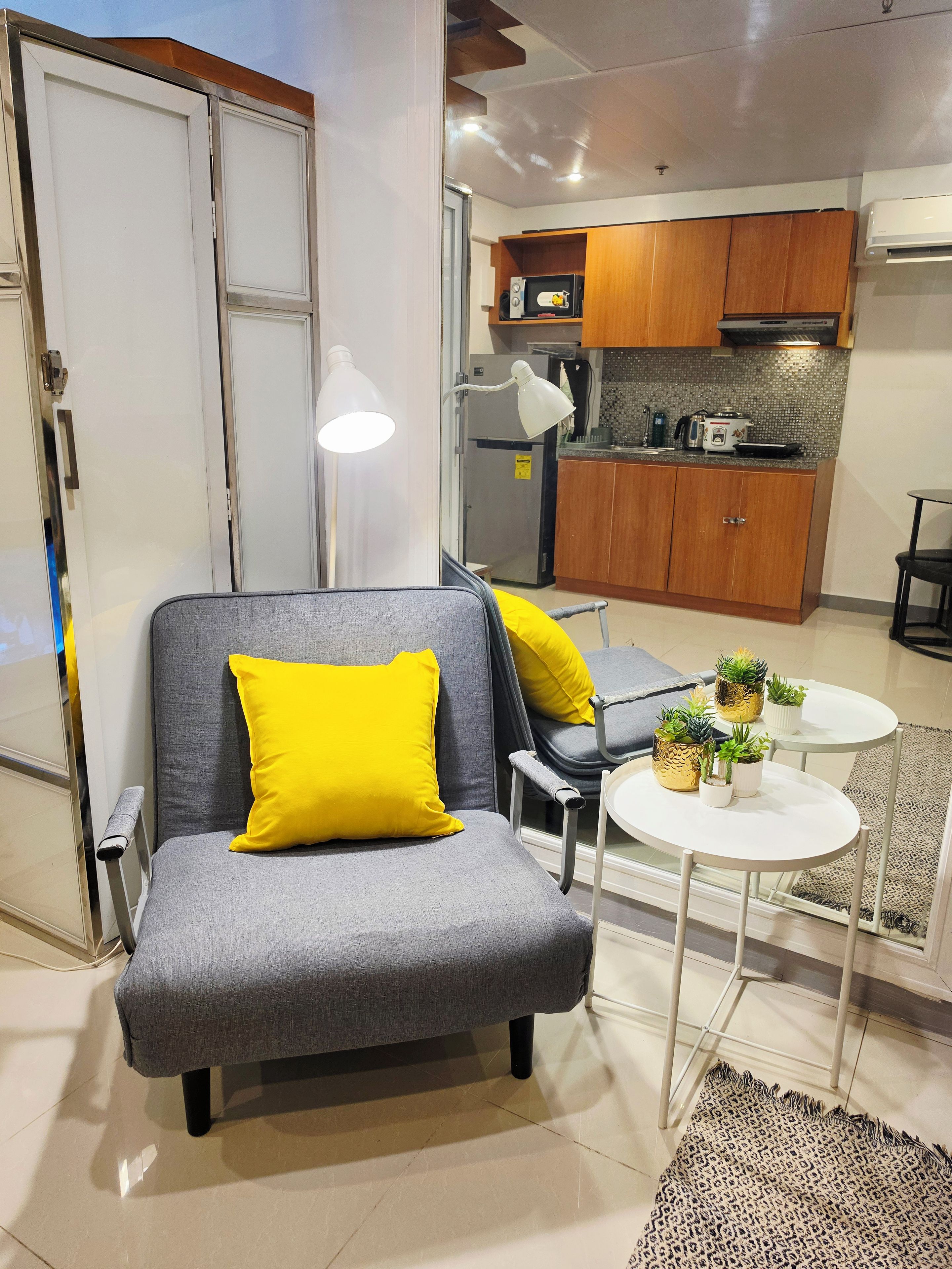 A modern living room with a grey sofa, yellow cushions, a small white table with potted plants, and a kitchen in the background.