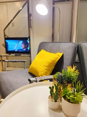 A modern living room with a television, a sofa with a yellow pillow, and two potted plants on a white table. - Casa Rosa2 2BR Loft in BGC With 55" Smart TV & 5 pax (Taguig)