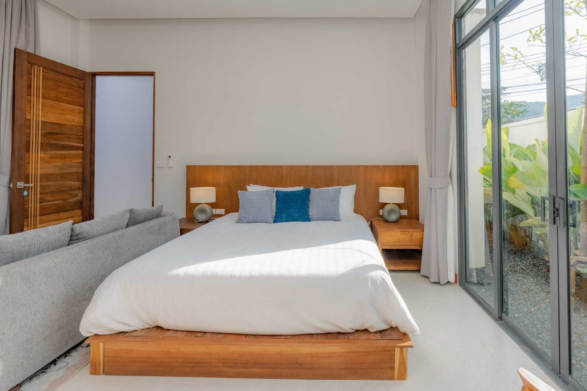 Ein modernes Schlafzimmer mit einem hölzernen Bett, einem grauen Sofa und einer Glastür, die auf einen Balkon führt.