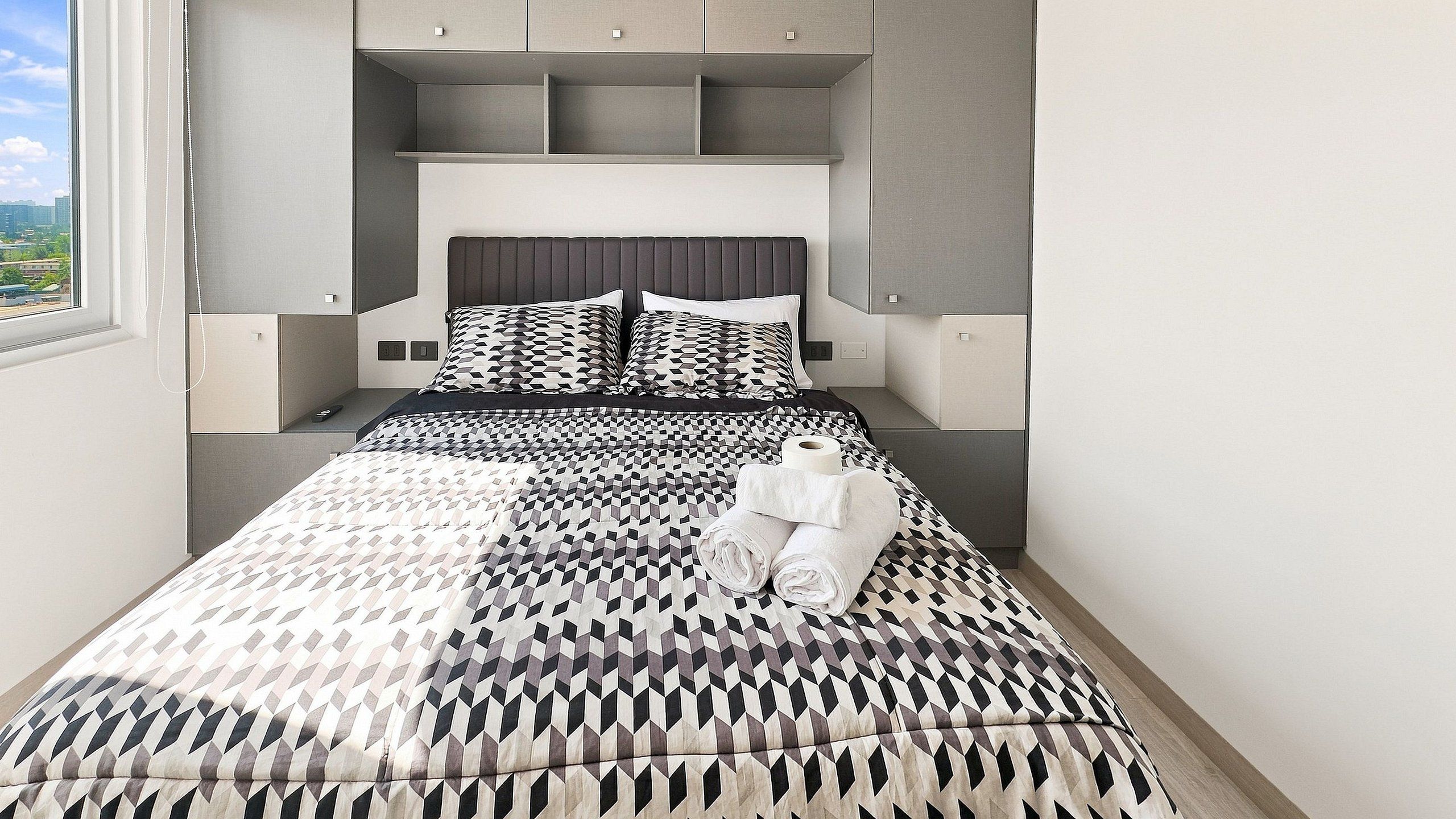 A modern bedroom with a geometric patterned bedspread, a neatly made bed with rolled towels, and built-in storage cabinets.