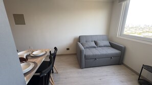 A room with a dining table, chairs, a grey sofa, a window with a city view, and a small cabinet. - Oceana Suites Ñuñoa Central (Santiago)