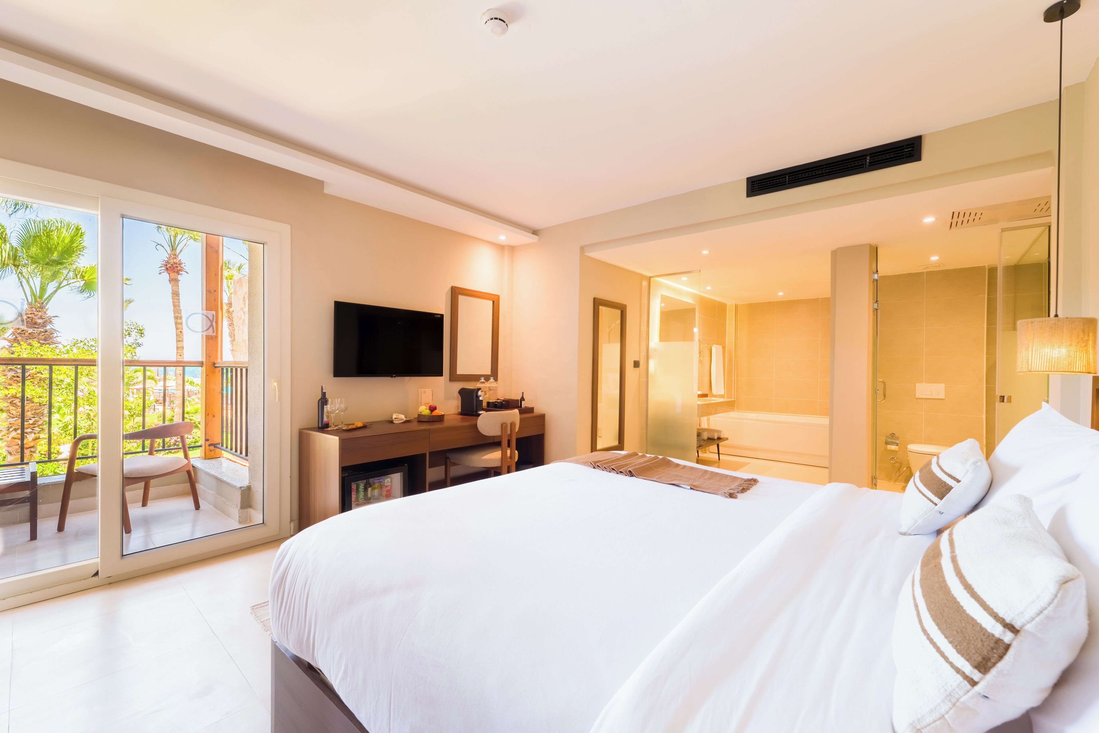 A hotel room with a bed, a desk with a TV, a bathroom, and a balcony with a chair.