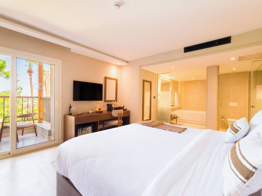 A hotel room with a bed, a desk with a TV, a bathroom, and a balcony with a chair.