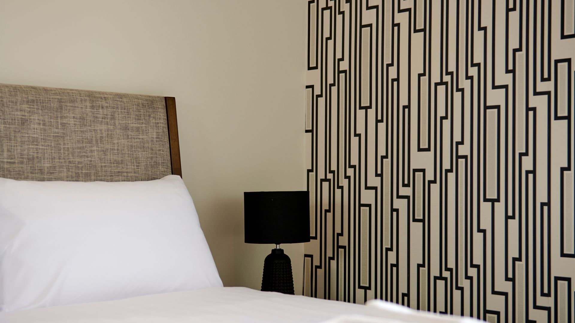 A hotel room with a bed, a headboard, a nightstand with a lamp, and a patterned wall.
