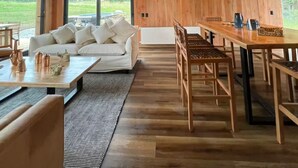 A spacious room with a wooden ceiling, a large window, a white sofa, a dining table, and chairs.