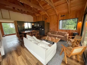 Un salón espacioso con un sofá, sillas y una mesa de centro. Se ve una cocina al fondo, y ventanas amplias ofrecen vistas al exterior.